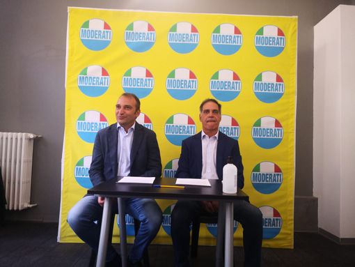 Due persone sedute a un tavolo con dietro bandiere di partito